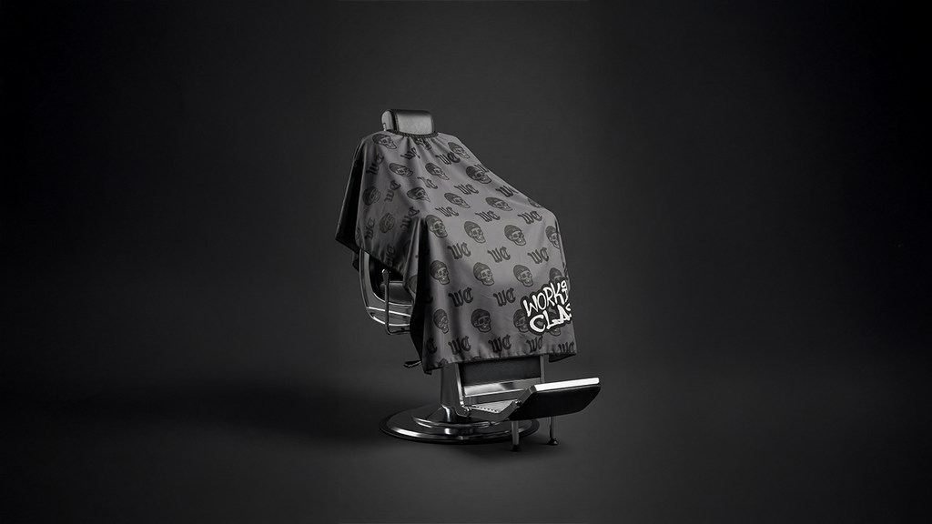 Monogram barber cape draped over a salon chair, featuring dual-closure neck system and durable fabric for professional hairdressing use.